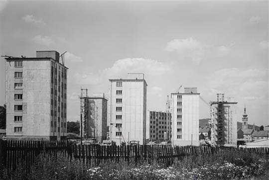 Černobílá fotografie rozestavěných panelových věžáků za dřevěným plotem, vpravo v pozadí věž písecké radnice