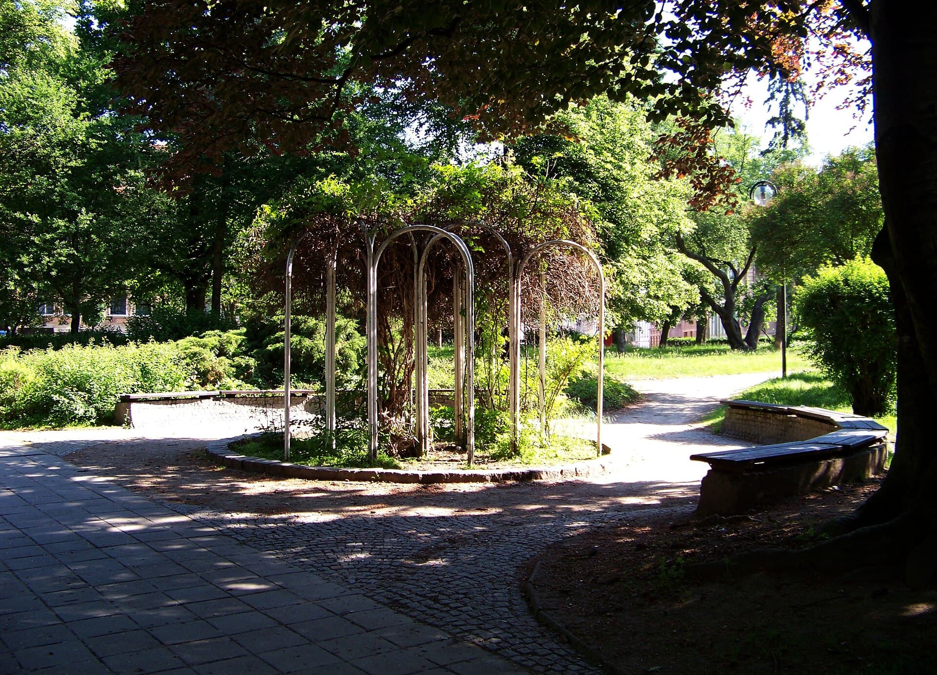 Park na Husově náměstí v Písku s kovovou pergolou porostlou popínavými rostlinami, dlážděnou cestou a lavičkami ve stínu stromů