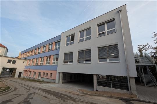 Nová třípodlažní šedobílá přístavba se dvěma patry na sloupech, pod ní kryté parkování, vlevo starší budova školy s modrými pásy oken
