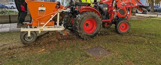 Malý červený traktor s oranžovou násypkou na cibulky projíždí trávníkem pod podzimními stromy