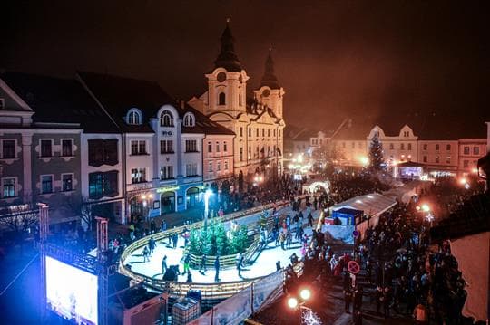Noční pohled shora na osvětlené kluziště s bruslaři na Velkém náměstí v Písku, vedle vánoční strom, stánky a kostel s dvěma věžemi