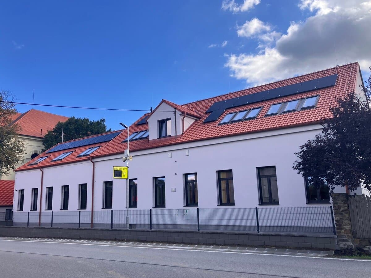 Bílá dvoupodlažní budova azylového domu ve Veselíčku po rekonstrukci, s novou červenou střechou, solárními panely, střešními okny a plotem podél silnice