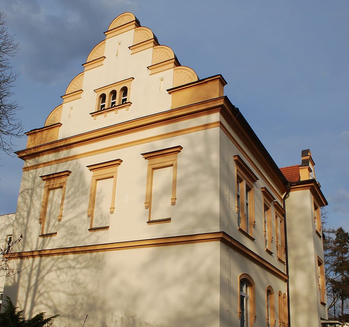 Světlá historická budova v areálu písecké nemocnice se stupňovitým štítem a okrovými šambránami, kolem stromy a keře