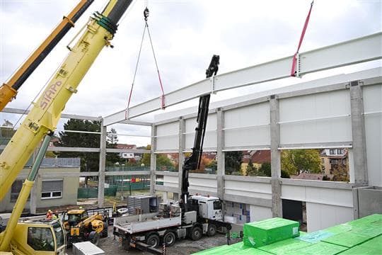 Dva autojeřáby zvedají ocelový vazník nad rozestavěnou halu s betonovými sloupy a světlým opláštěním