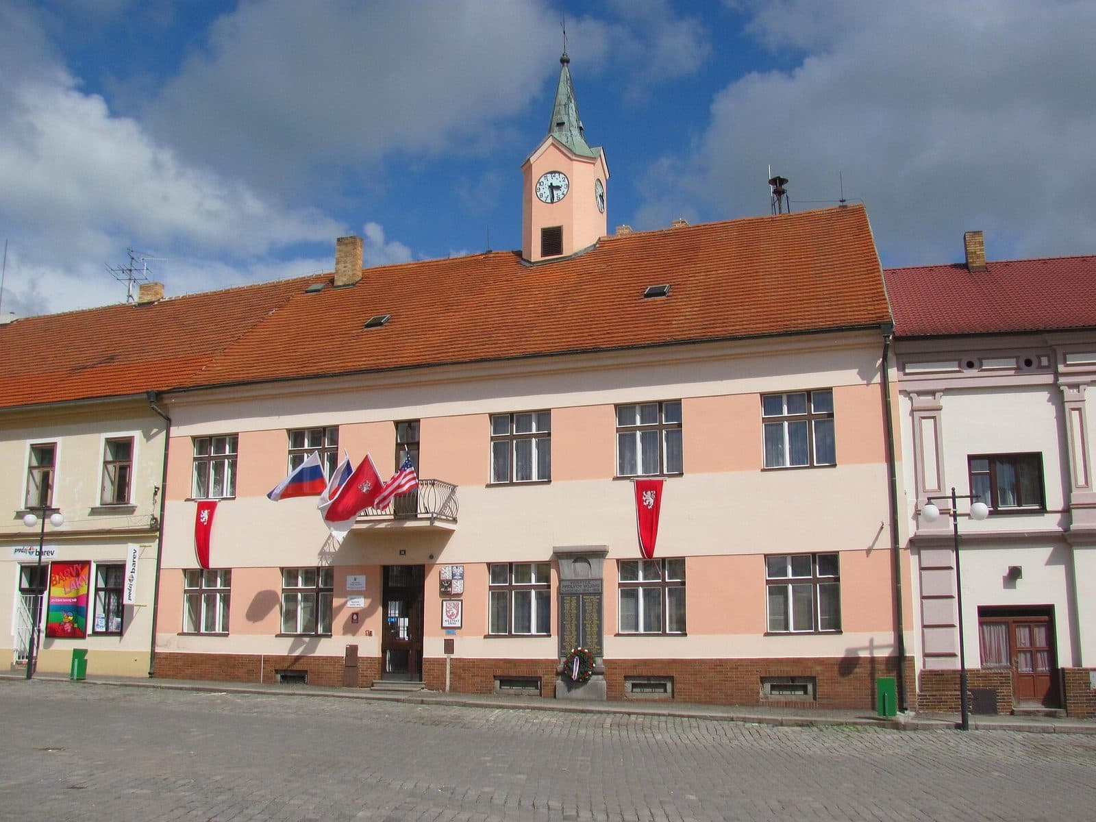 Budova městského úřadu v Mirovicích s hodinovou věžičkou, vlajkami na balkoně a pamětní deskou na fasádě, před ní dlážděné náměstí